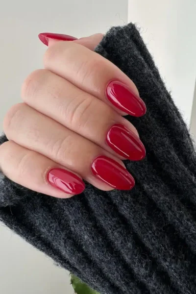 Manicura Roja Clásica Brillante y Elegante Mano con uñas largas ovaladas pintadas de un vibrante color rojo clásico brillante, sujetando una manga de jersey gris oscuro.